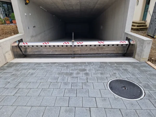 Eine mobile Hochwasserschutzwand sperrt den Zugang zur Tiefgarage, davor graue Pflastersteine und ein runder Schachtdeckel.