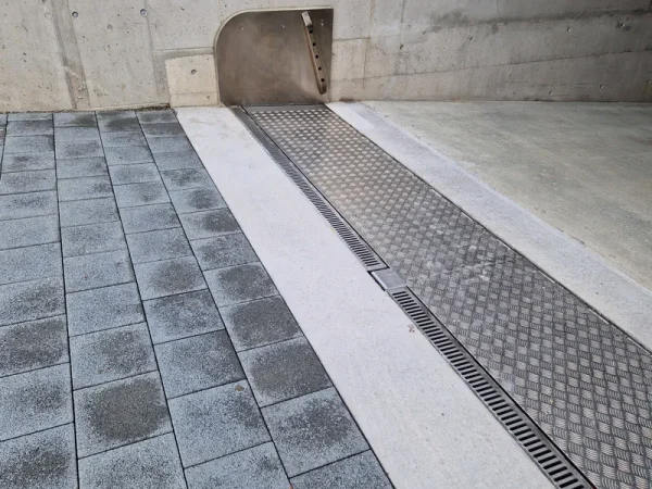 Detailansicht eines modernen Entwässerungssystems zwischen Beton, Pflastersteinen und Rinnengitter an einer glatten Betonwand.