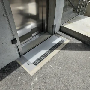 Eine Lifttür öffnet sich auf Bodenhöhe mit Metallübergang und Entwässerungsrinne, umgeben von Beton und Treppe rechts.
