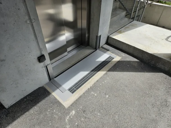 Eine Lifttür öffnet sich auf Bodenhöhe mit Metallübergang und Entwässerungsrinne, umgeben von Beton und Treppe rechts.