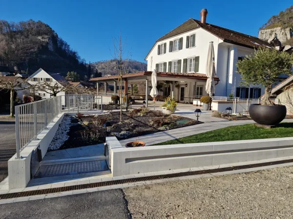 Großes weißes Haus mit grünen Fensterläden, braunem Dach und Hochwasserschutzrampe im Garten vor Hügeln unter blauem Himmel.
