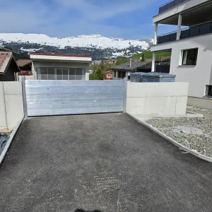 Ein Metallschott verschliesst die neu asphaltierte Zufahrt, umgeben von Betonschutzwänden, modernen Gebäuden und schneebedeckten Bergen.