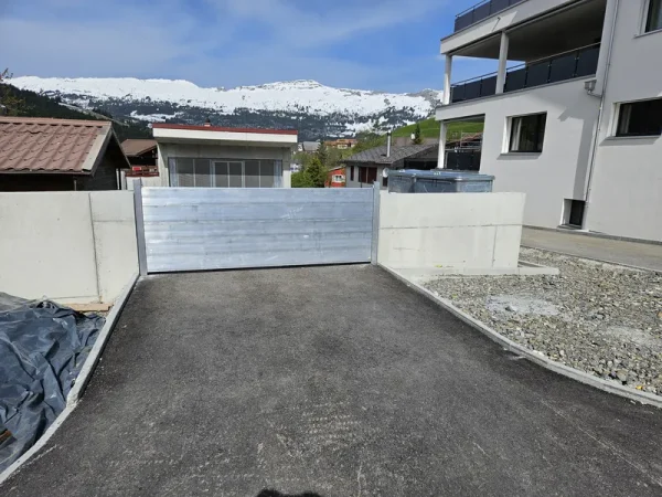 Ein Metallschott verschliesst die neu asphaltierte Zufahrt, umgeben von Betonschutzwänden, modernen Gebäuden und schneebedeckten Bergen.