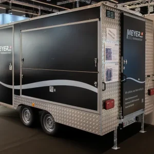 Schwarzer und silberner MEYER-Doppelachs-Anhänger mit Metallverkleidung, Seitentür und Infotafeln zum Hochwasserschutz.