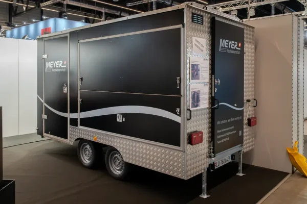 Schwarzer und silberner MEYER-Doppelachs-Anhänger mit Metallverkleidung, Seitentür und Infotafeln zum Hochwasserschutz.