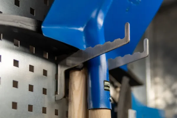 Nahaufnahme eines Metallhakens, der eine blaue Schaufel und ein Holzwerkzeug an einer Lochwand im Hochwasserschutz-Lager hält.