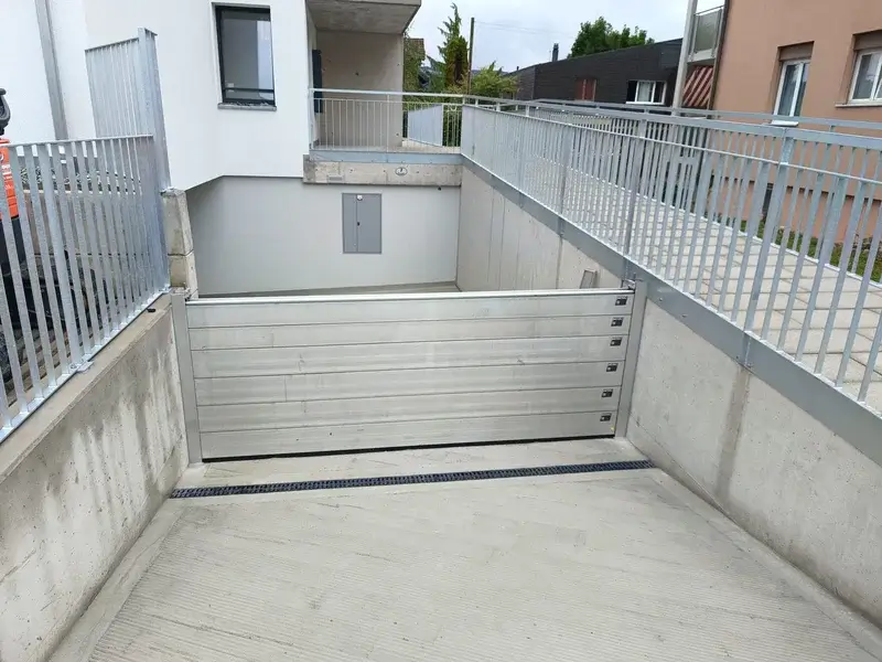 Ein Metallsicherheitstor mit Hochwasserschutz sperrt eine Tiefgarageneinfahrt bei modernen Wohnhäusern, umgeben von Beton.