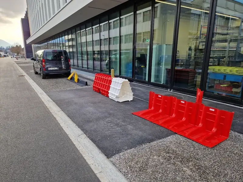 Trottoir mit rot-weissen mobilen Hochwasserschutzbarrieren vor Glasgebäude; schwarzer Lieferwagen daneben geparkt.