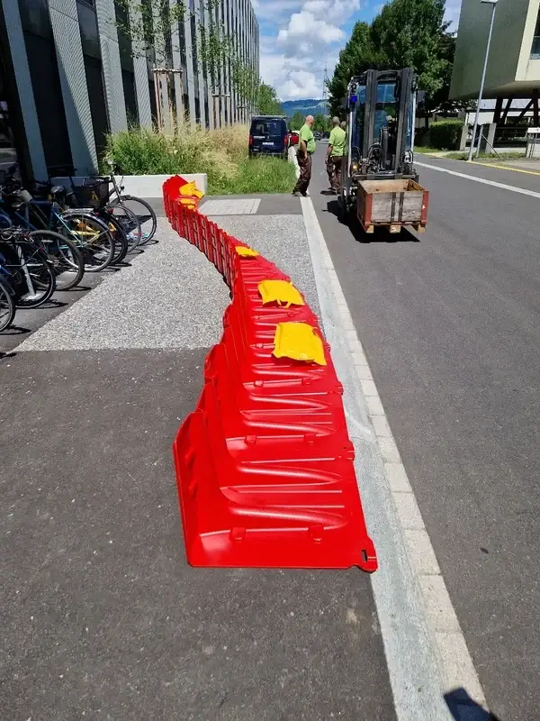 Rote Kunststoff-Hochwasserschutzbarrieren mit gelben Abdeckungen, Gabelstapler, Velos und Gebäude unter blauem Himmel.