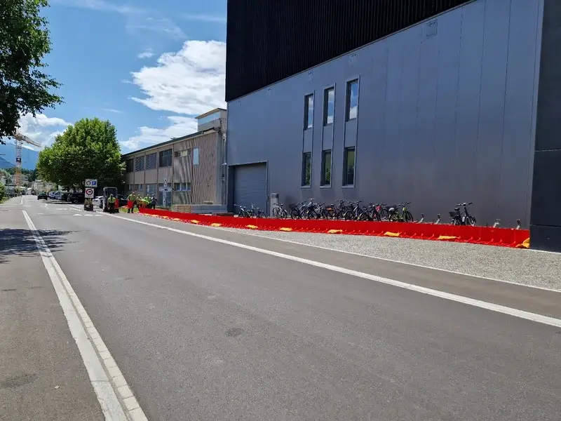 Eine Reihe rot-gelber Hochwasserschutzbarrieren steht am Trottoir neben einem Gebäude mit abgestellten Velos, Strasse leer.
