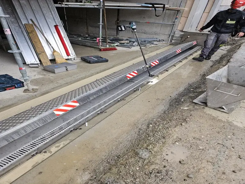 Baustelle mit eingebaurem Metall-Entwaesserungskanal, rot-weiss markiert, Werkzeug, Leitern und Arbeiter mit Helm im Hintergrund.