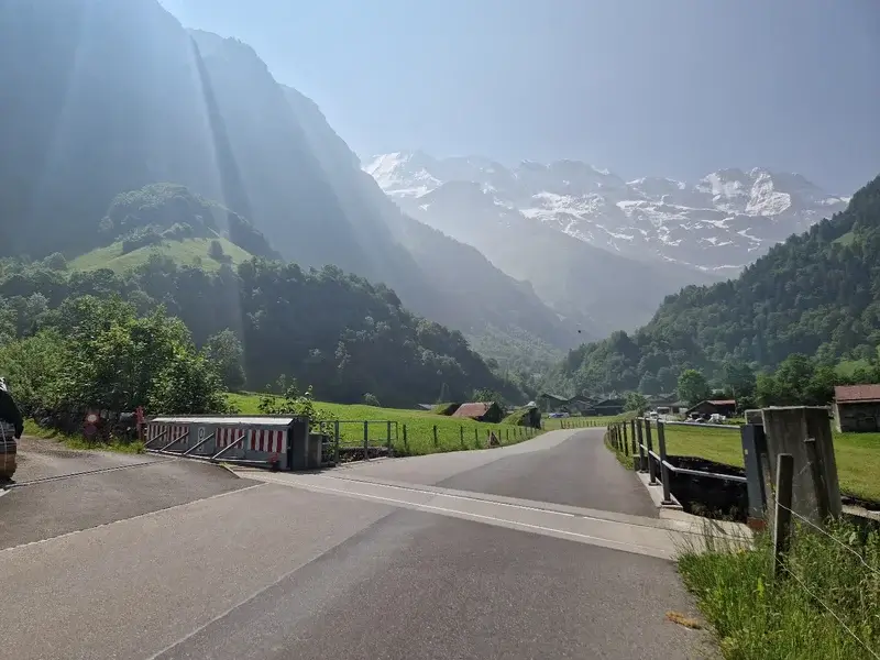 Eine asphaltierte Straße führt durch ein grünes Tal mit schneebedeckten Bergen, Sonnenstrahlen und verstreuten Häusern.