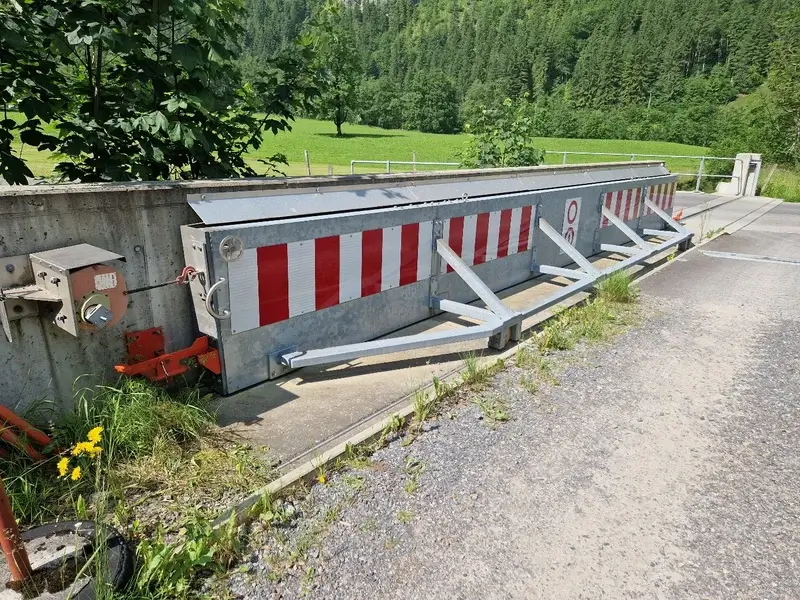 Eine rot-weiss gestreifte Hochwasserschutzbarriere sperrt eine Nebenstrasse im Grünen, verstärkt mit Schrägstützen.
